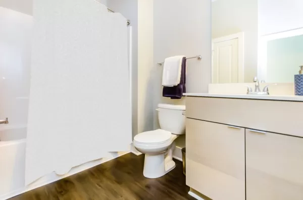 Bathroom with wooden floors, beige cabinets, and white appliances, including a toilet, shower/bath, and sink